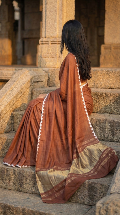 Woman wearing a chocolate-brown-modal-silk-saree-lagdi-patta-pallu, sitting on concrete chairs in an architectural setup.