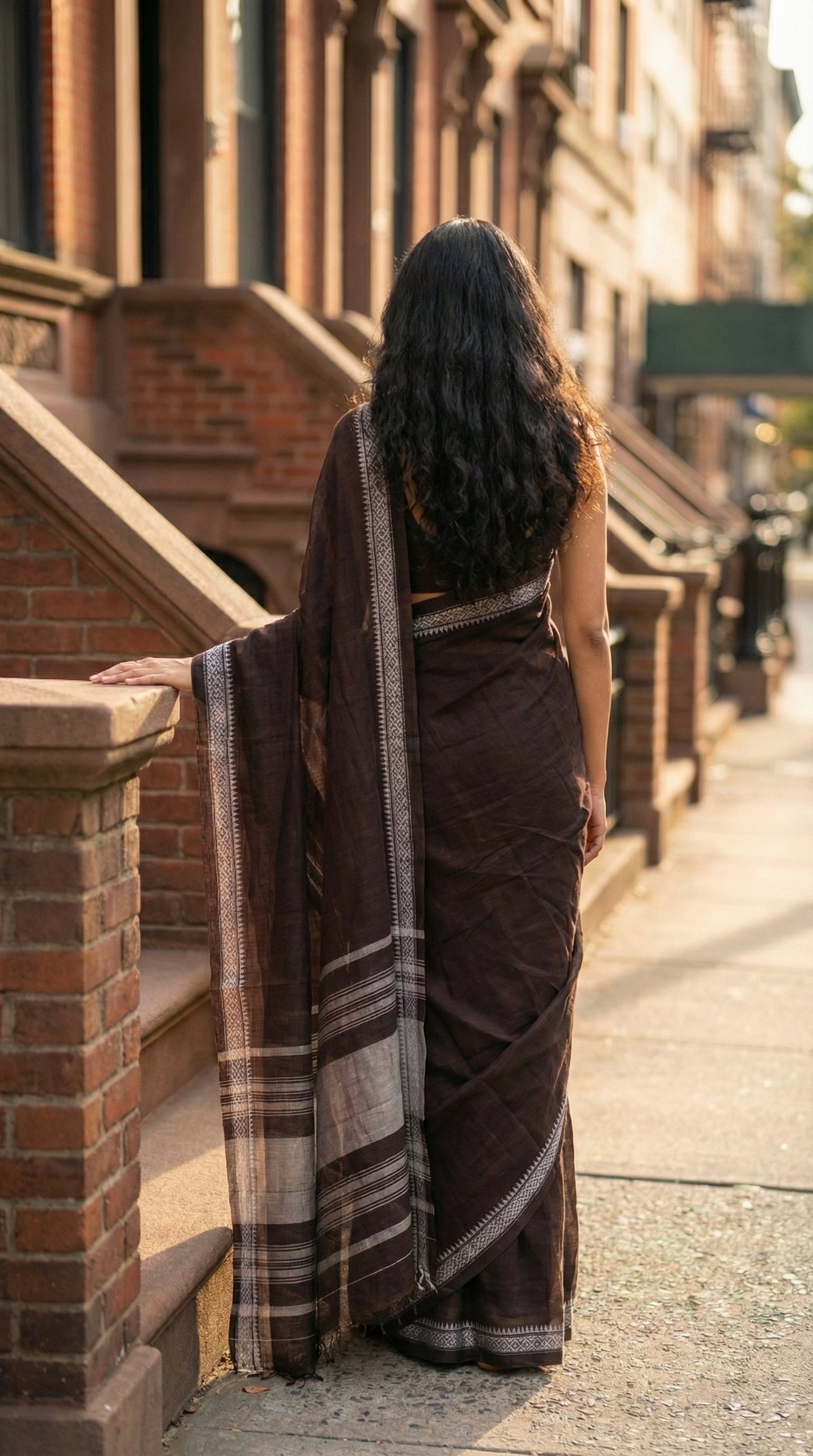 Woman in a chocolate-brown-mul-cotton-saree, standing on a city street with brick buildings.