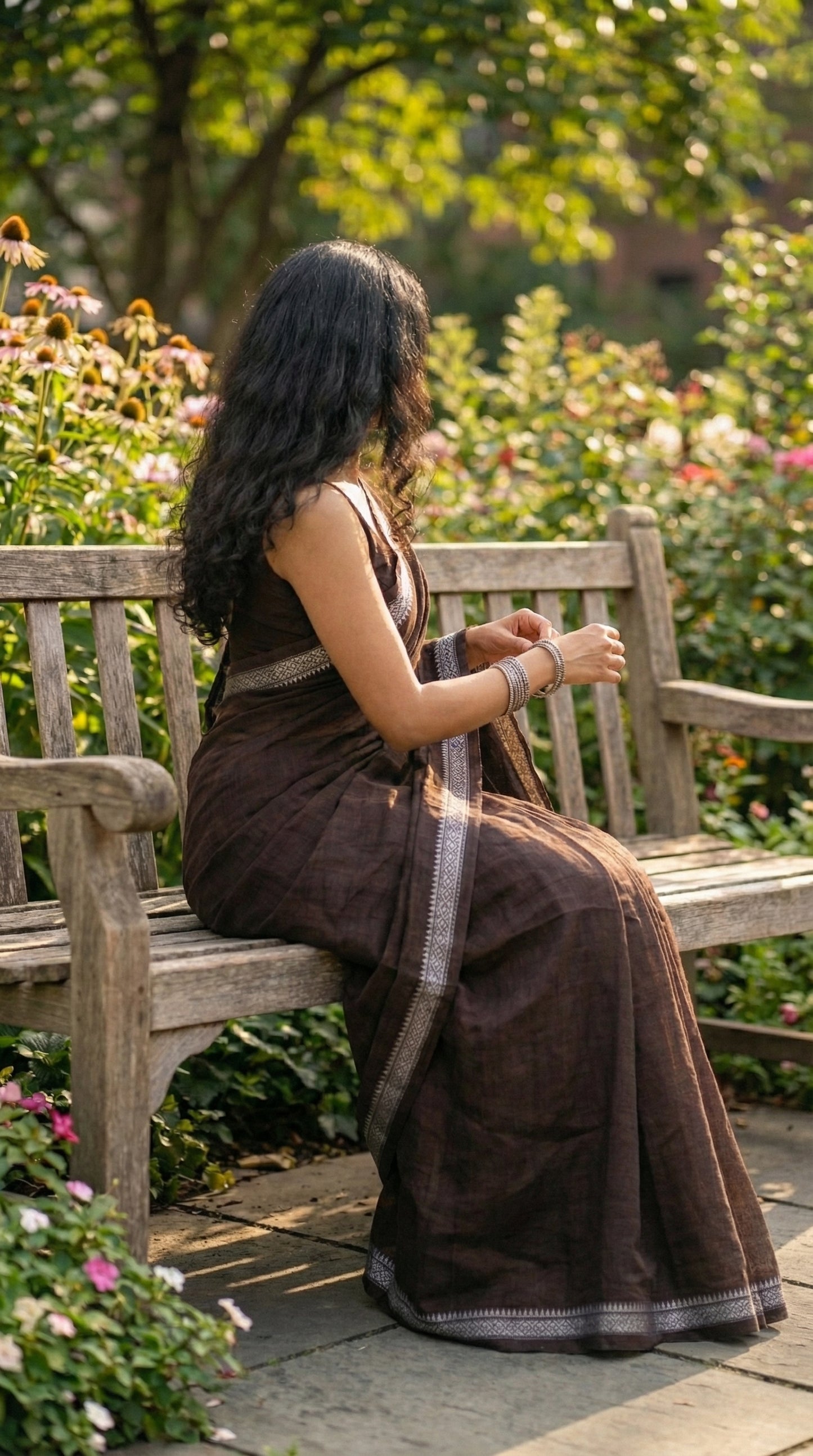 Woman in a chocolate-brown-mul-cotton-saree, sitting on a wooden bench in a garden.