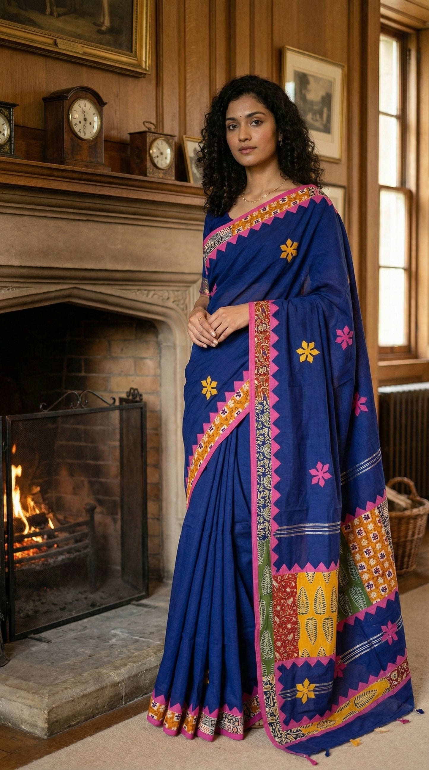 Woman wearing a blue cotton saree with multicolor patchwork and floral embroidery, in a room with a fireplace.