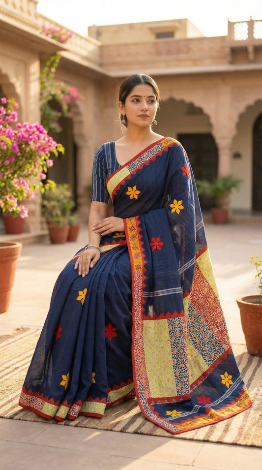 Woman wearing a navy blue cotton saree featuring vibrant appliqué panels and zigzag borders, artisan-made ethnic wear in an outdoor setting with architectural elements and plants.