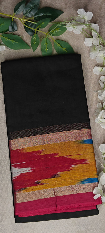 Black cotton saree with vibrant Ikat-style border in red, gold, and blue, folded with floral accents with neutral background.