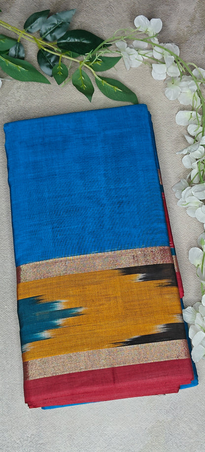 Blue cotton saree with mustard and black Ikat border and red edge, displayed with white flowers and neutral background.
