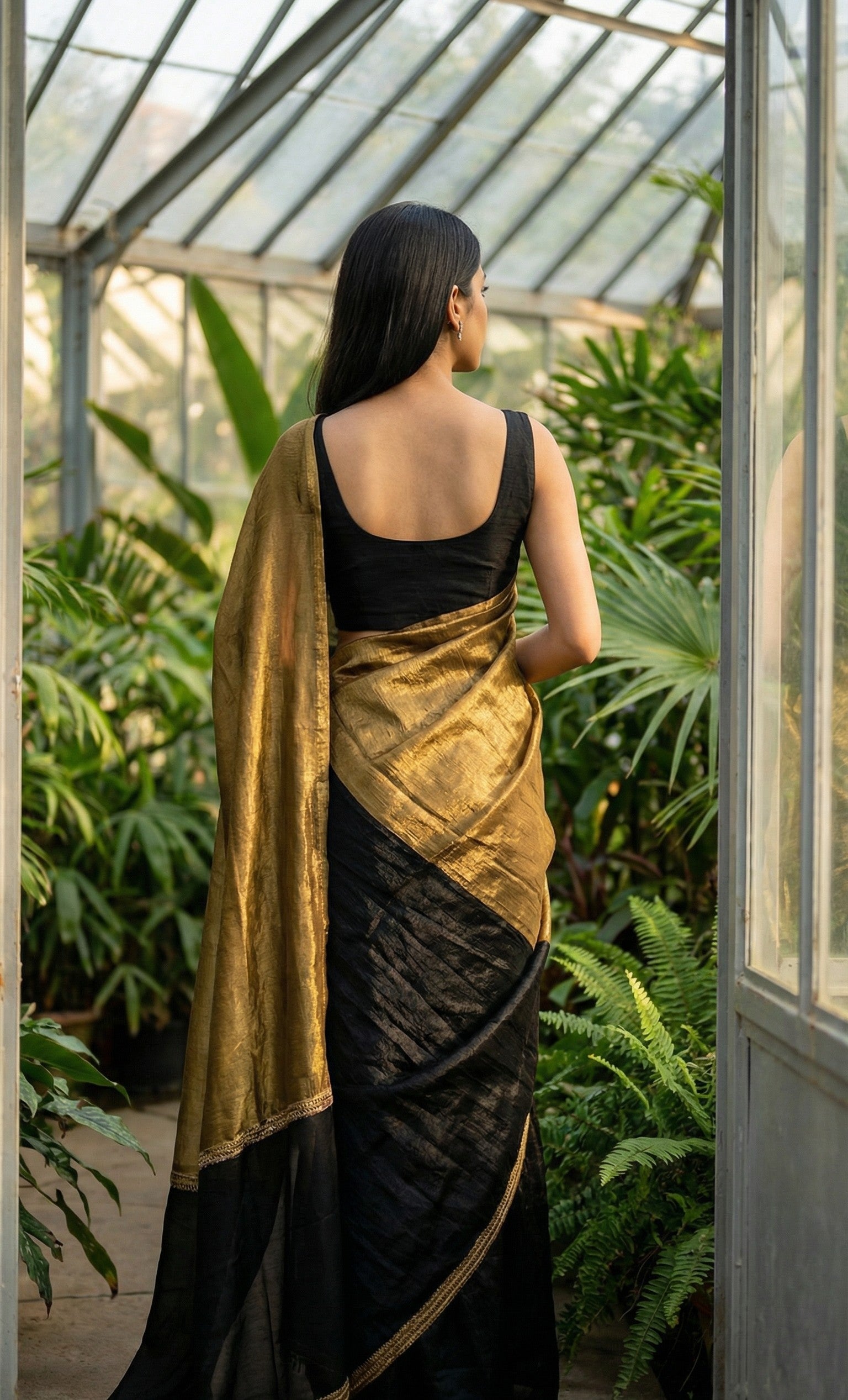 Woman in a antique golden Fendy silk saree with floral motifs and black contrast border, elegant festive wear, standing in a greenhouse filled with plants.