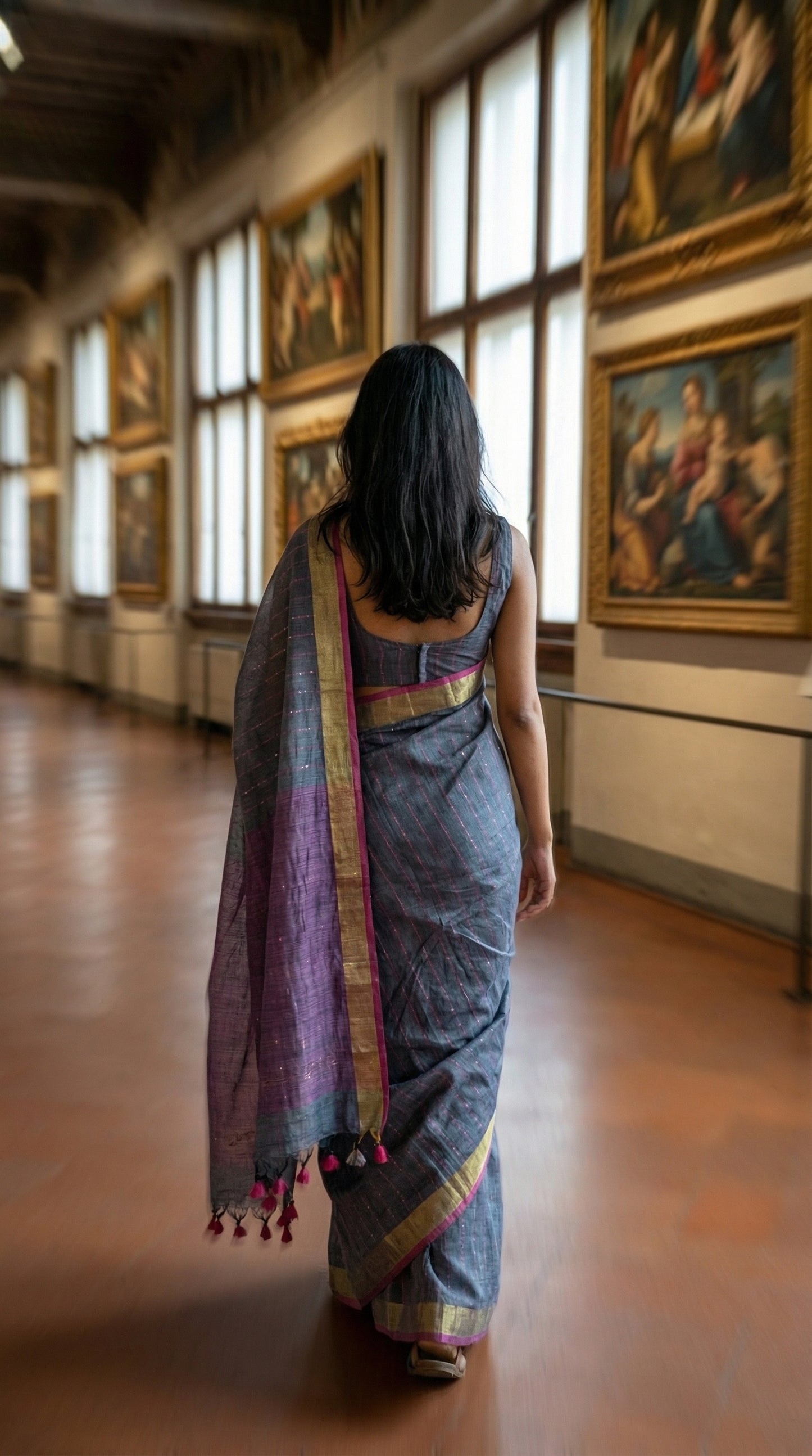 Woman in a grey-cotton-linen-saree, walking through a museum with paintings on the walls.
