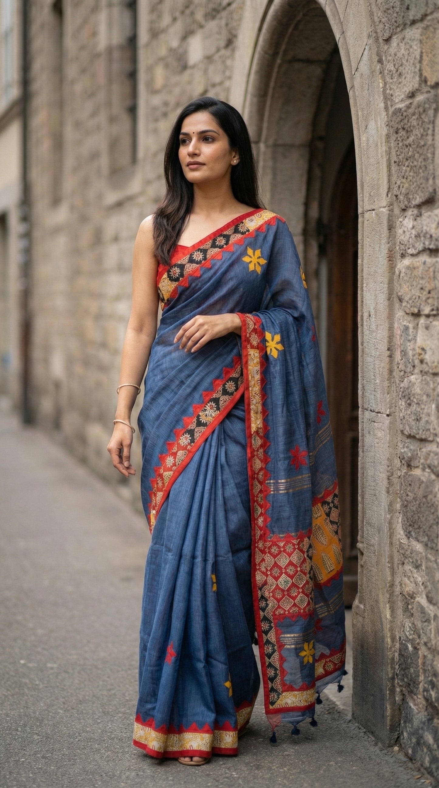 Woman in a grey linen cotton saree with traditional applique border, standing against a stone wall.