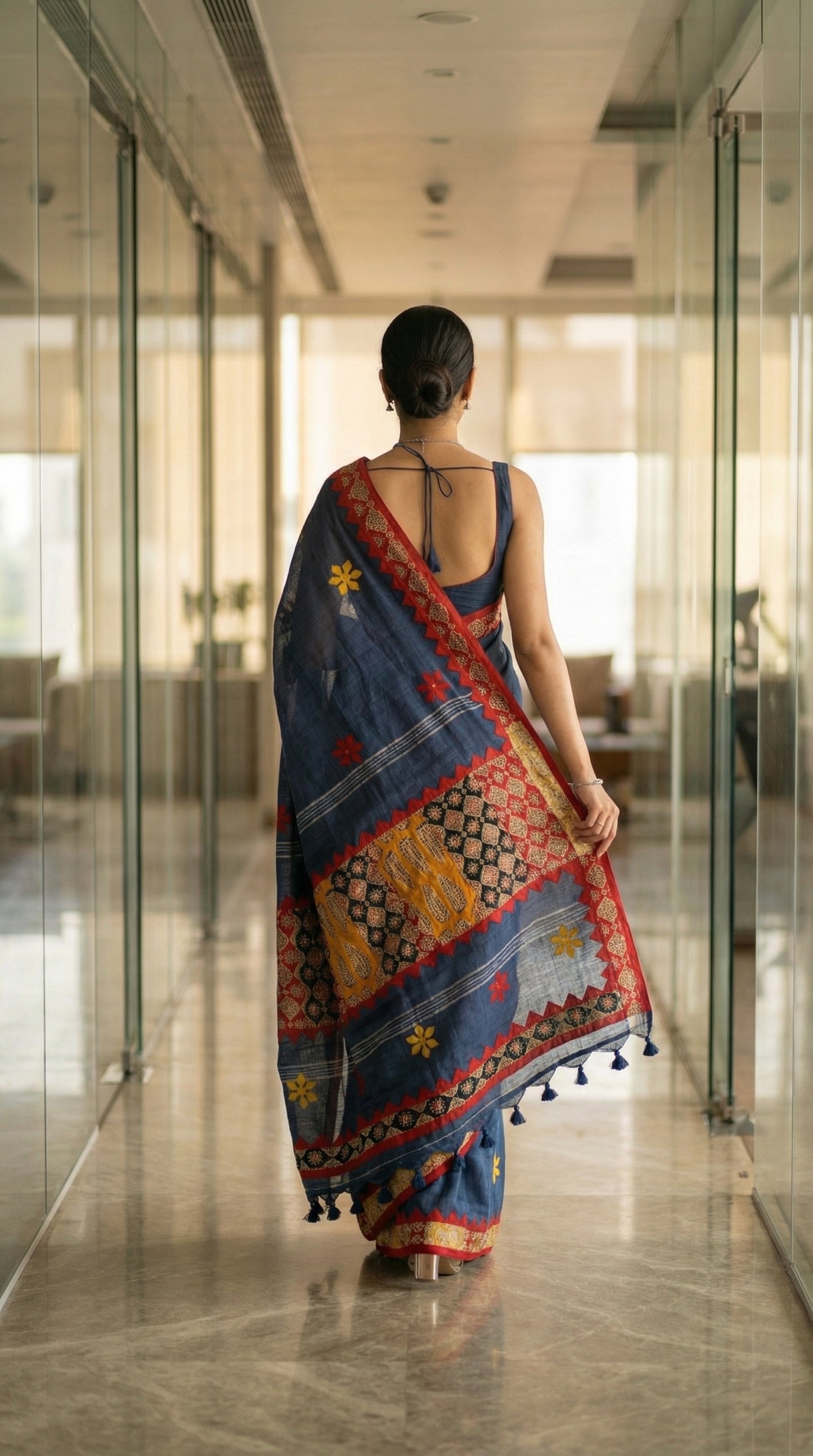 Woman in a grey linen cotton saree with handcrafted applique border, walking through a modern indoor setting.