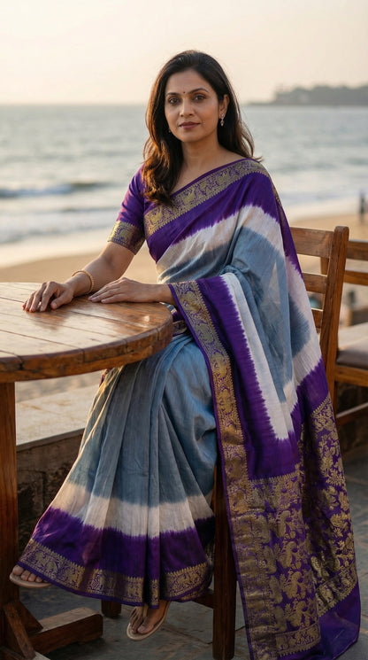 Woman wearing grey modal silk saree with purple Zari border, sitting by a waterfront.