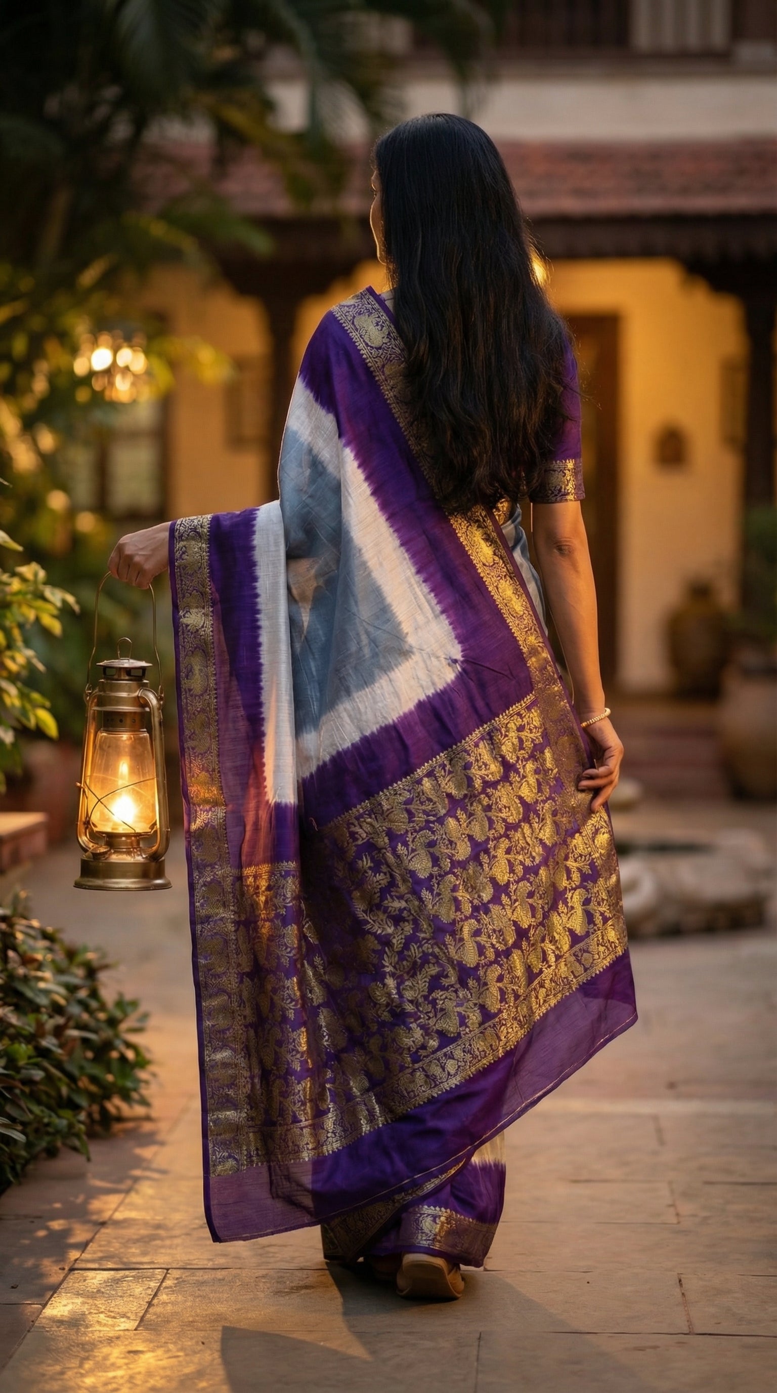 Woman wearing grey modal silk saree with purple Zari border, holding a lantern, standing on a stone path.