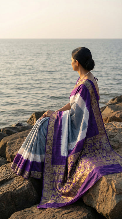 Woman wearing grey modal silk saree with purple Zari border, sitting on rocks by the ocean.