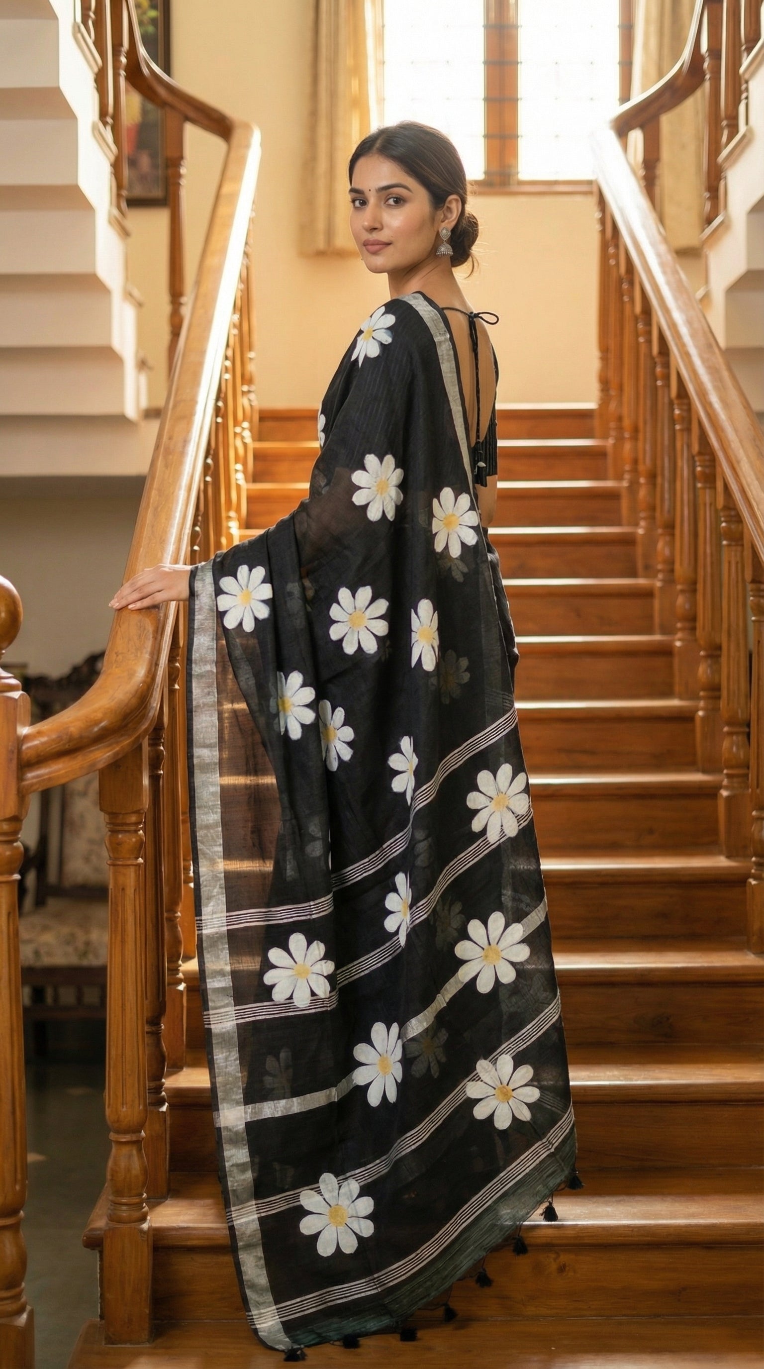 Woman wearing black cotton saree with bold white sunflower motifs and yellow centers, standing on staircase in her house.