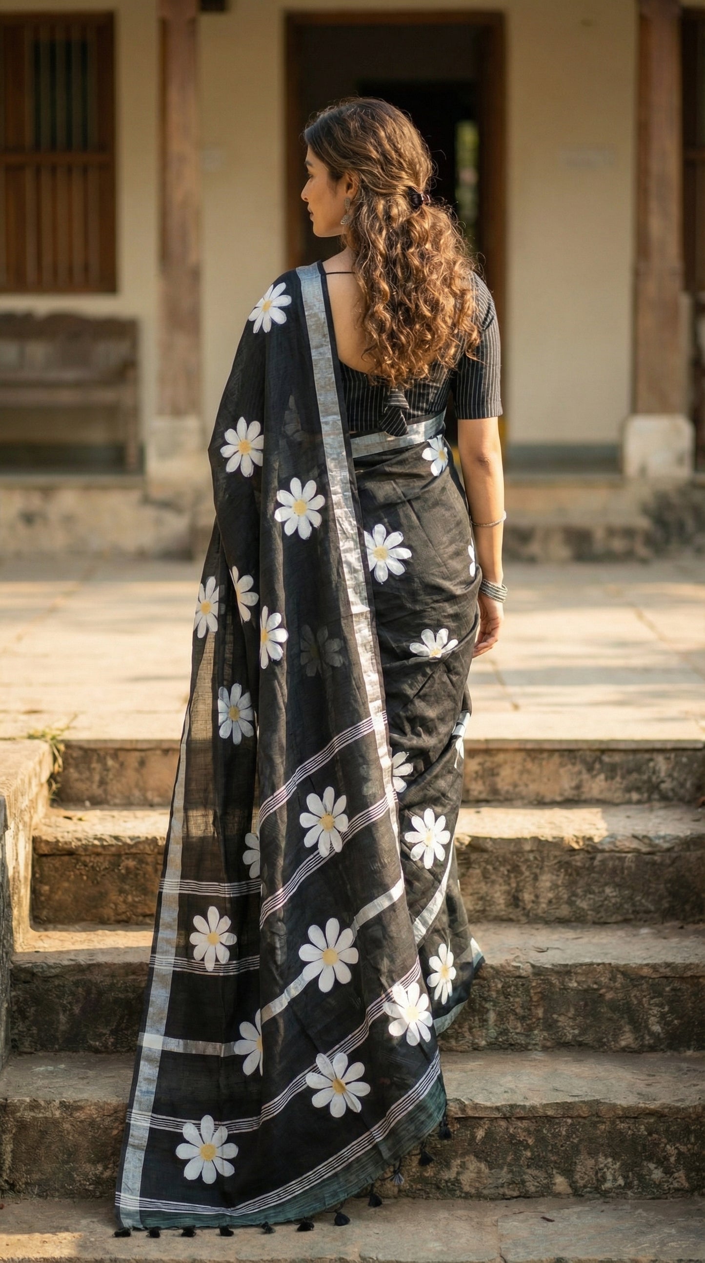 Woman wearing black cotton saree with bold white sunflower motifs and yellow centers, climbing stairs to enter a house.