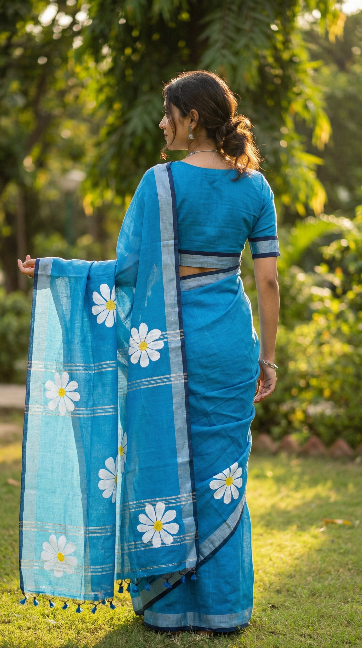 Woman wearing blue cotton saree with white and yellow sunflower motifs, hand-painted artisan saree for cultural occasions. Back to camera, pallu draped over forearm, head slightly turned; border + back silhouette visible.
