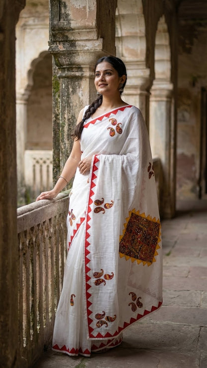 Woman in a white cotton saree with colorful hand-appliqué embroidery and red-yellow geometric border standing in an architectural setting.