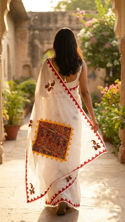 Woman in a white cotton saree with colorful hand-appliqué embroidery and red-yellow geometric border walking in a garden setting.