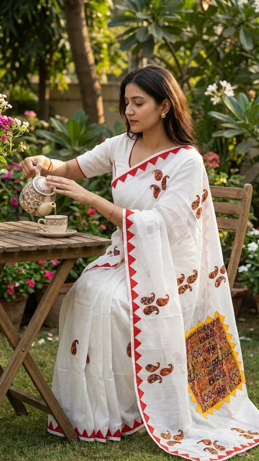 Woman in a white cotton saree with colorful hand-appliqué embroidery and red-yellow geometric border sitting outdoors, pouring tea.