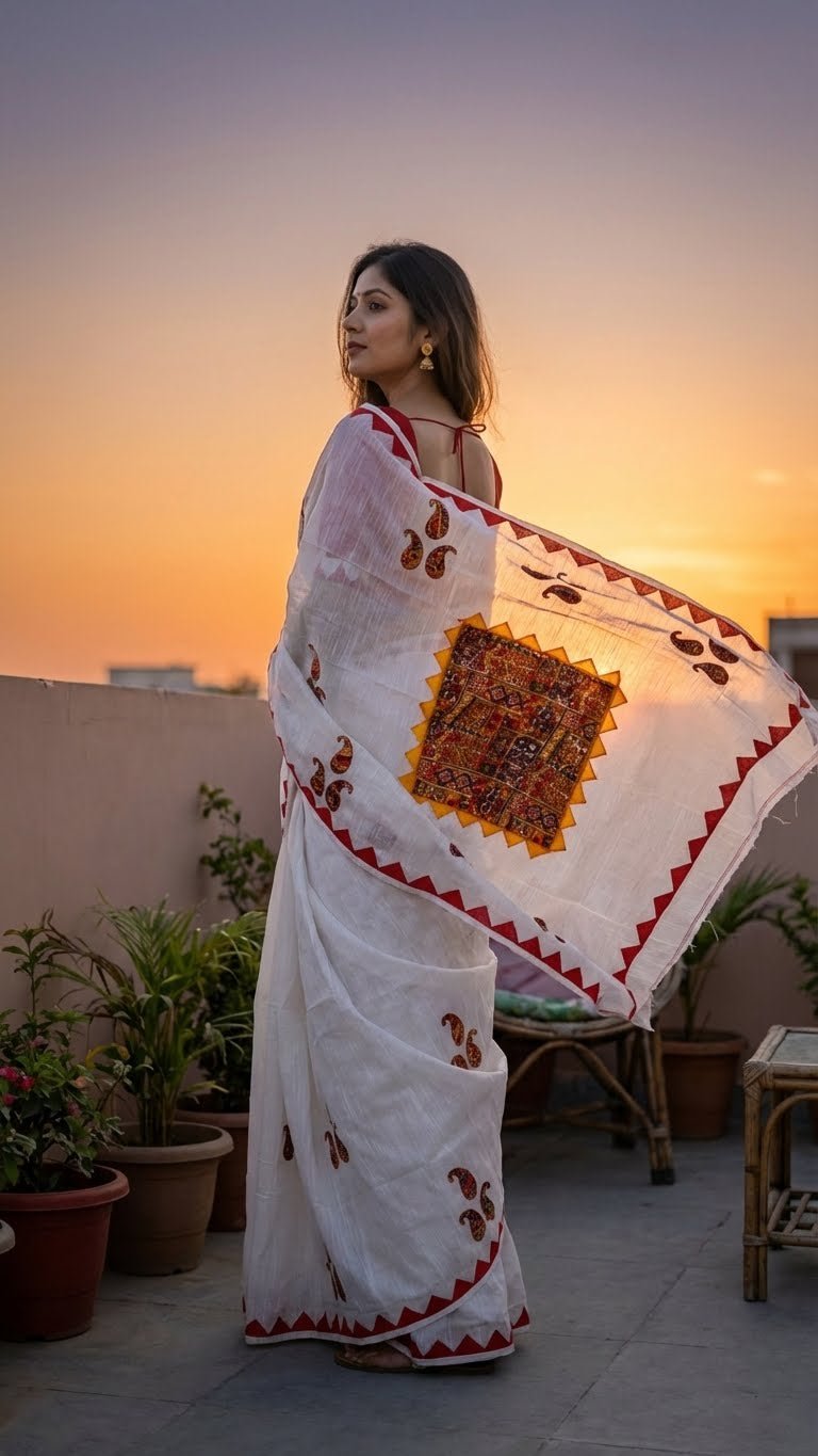 Woman in a white cotton saree with colorful hand-appliqué embroidery and red-yellow geometric border standing on a rooftop at sunset.