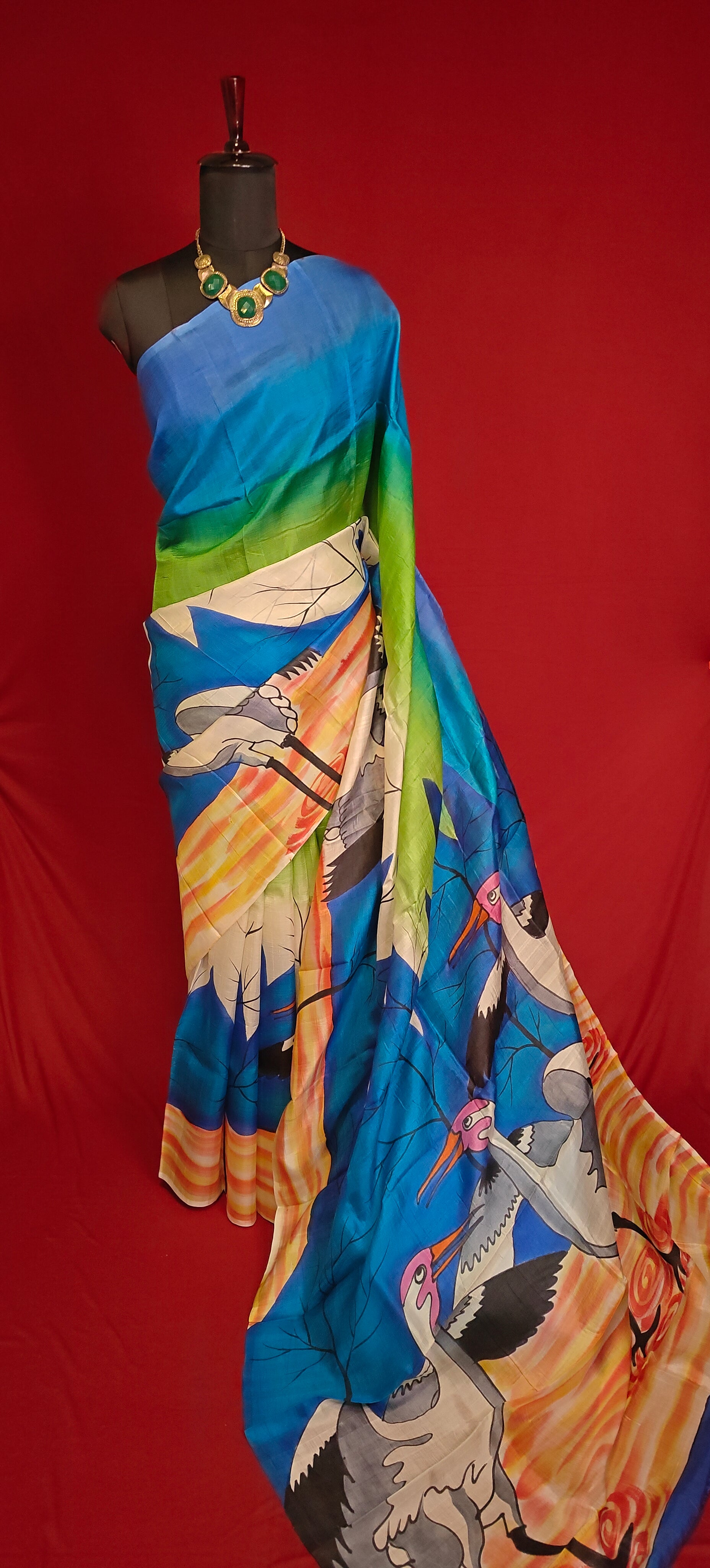 Hand-painted pure silk saree in dual-tone blue-green with crane motif and artisan detailing on a mannequin against a red background.