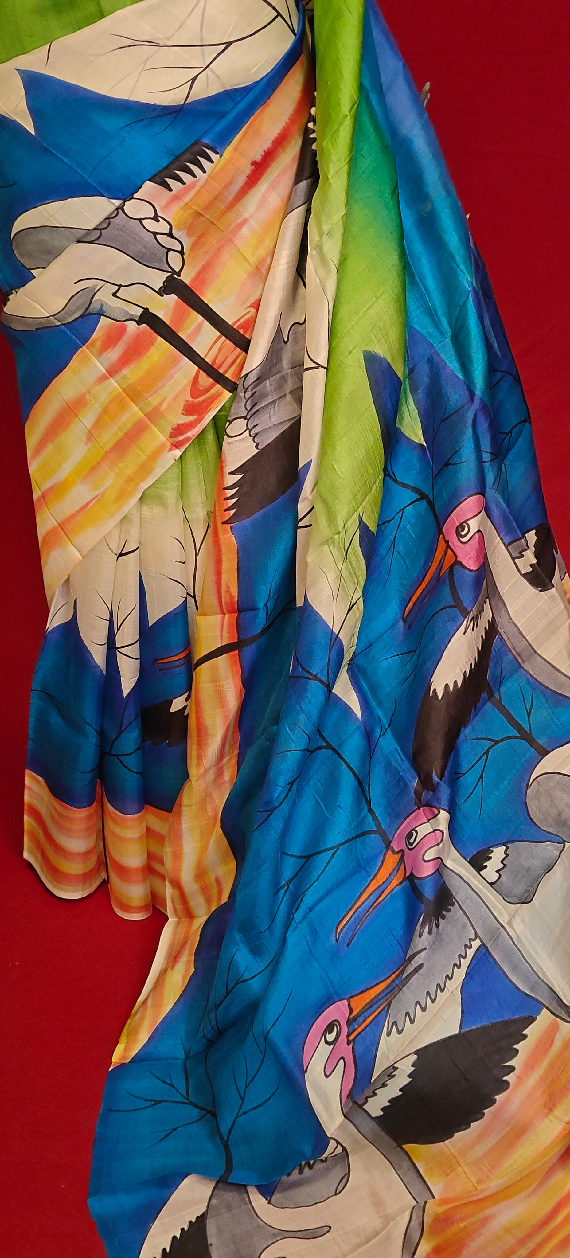 Hand-painted pure silk saree in dual-tone blue-green with crane motif and artisan detailing showcasing its artistic pallu against red background.