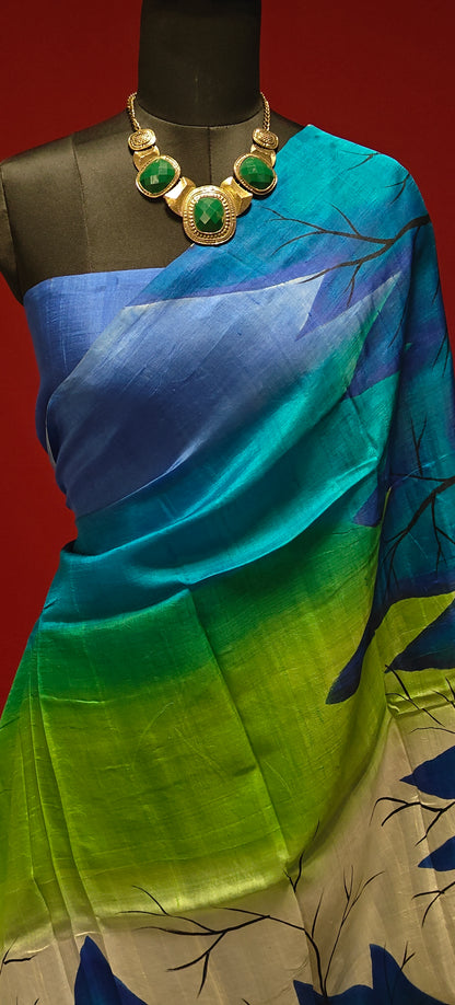 Hand-painted pure silk saree in dual-tone blue-green with crane motif and artisan detailing showcasing it's plain blue blouse piece draped on a mannequin against a red background.