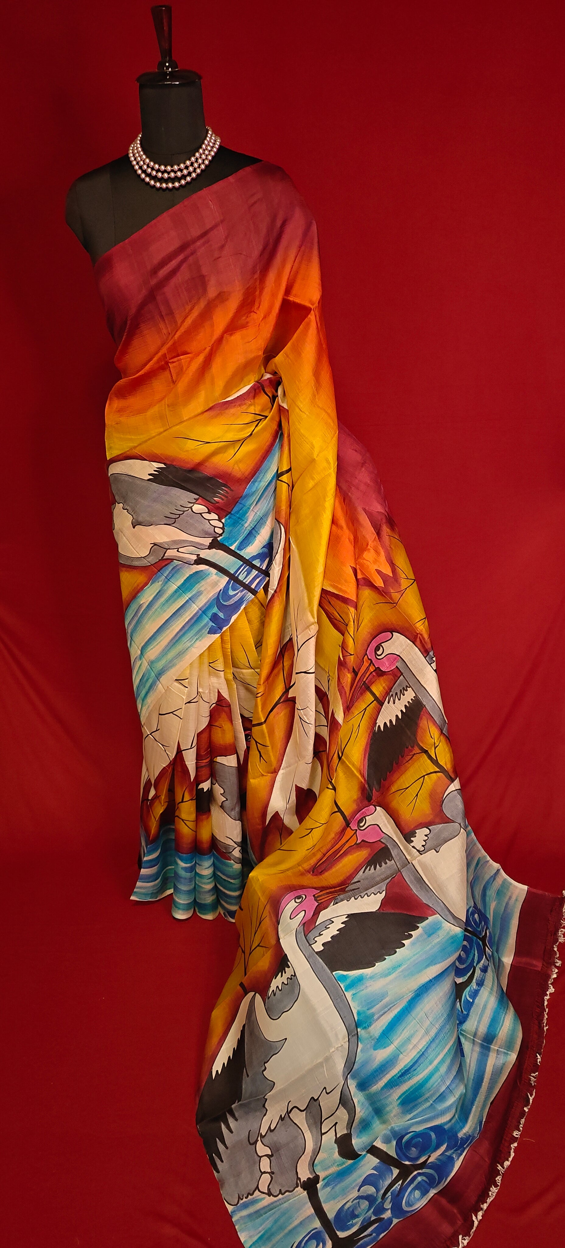 Hand-painted pure silk saree in dual-tone red and orange with crane motif and nature-inspired artwork on a mannequin against red background.