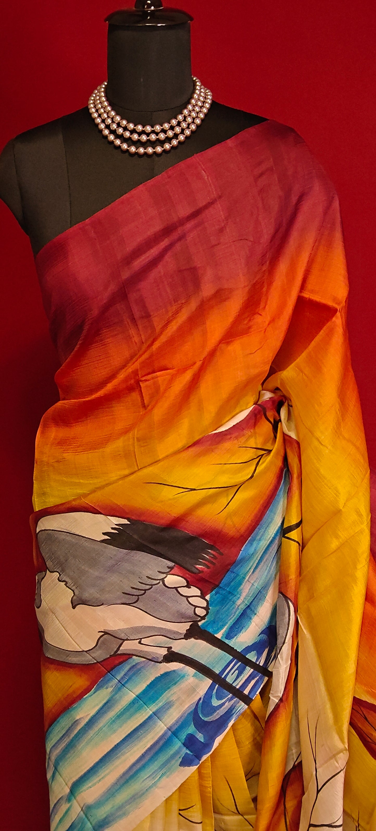 Hand-painted pure silk saree in dual-tone red and orange with crane motif and nature-inspired artwork on a mannequin against red background.