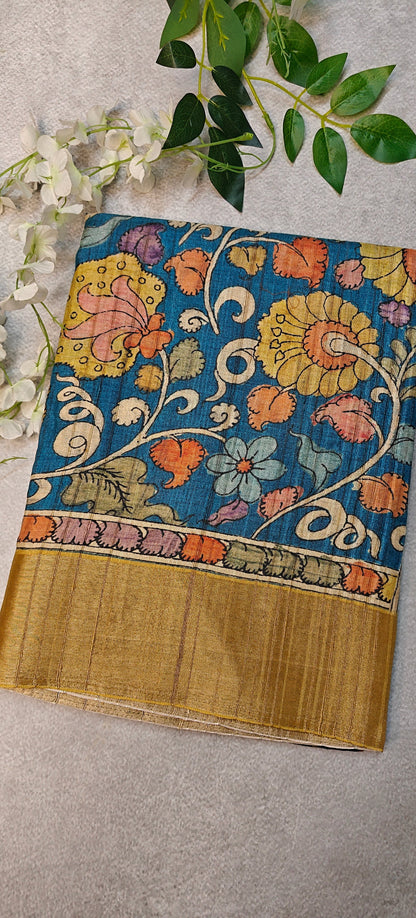 Blue saree with Kalamkari-inspired floral vine digital print and golden border lying flat and folded on a beige background with flowers.