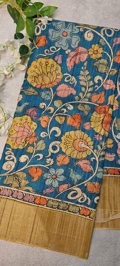 Blue saree with Kalamkari-inspired floral vine digital print and golden border lying flat and folded on a beige background with flowers.