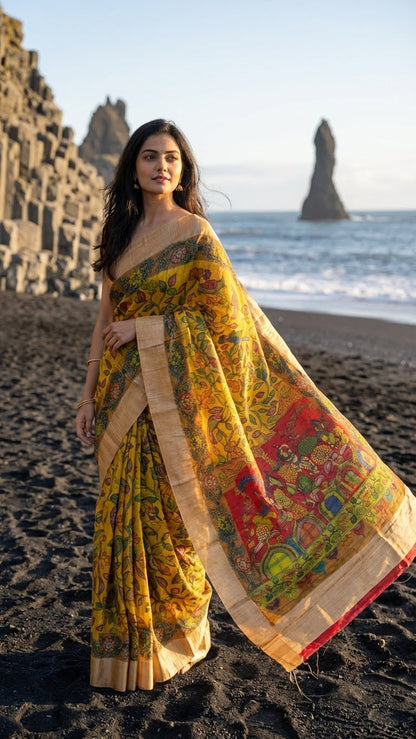 Woman in a yellow saree with kalamkari-style bird and floral digital print and golden border standing on a beach with ocean and rock formations in the background.