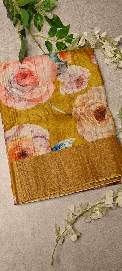 Golden yellow blended silk saree with Kalamkari-inspired floral digital print in pink, blue, and purple tones on a textured surface with greenery.
