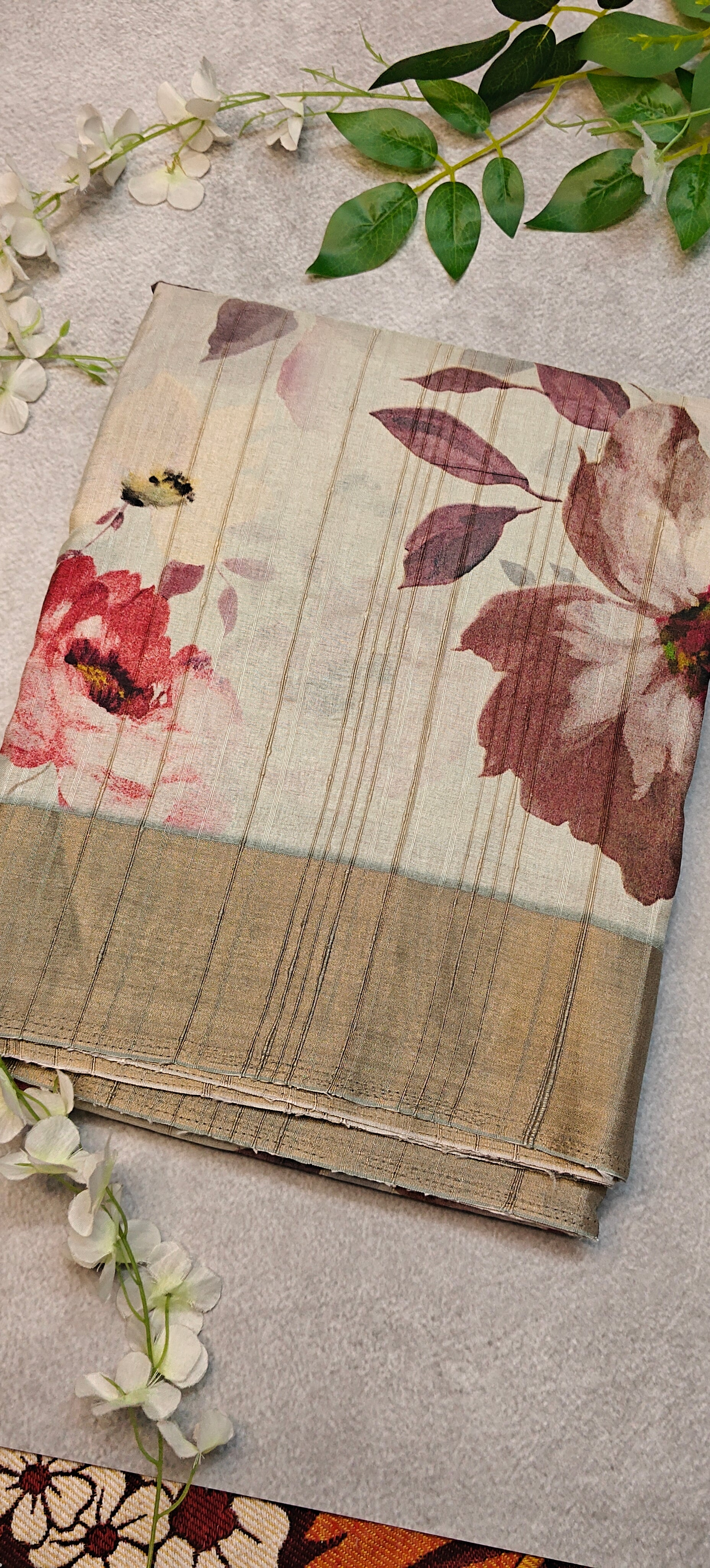 White blended silk saree with Kalamkari-inspired floral digital print in red, pink, and maroon tones with golden border lying flat and folded on a textured surface with green leaves and flowers.