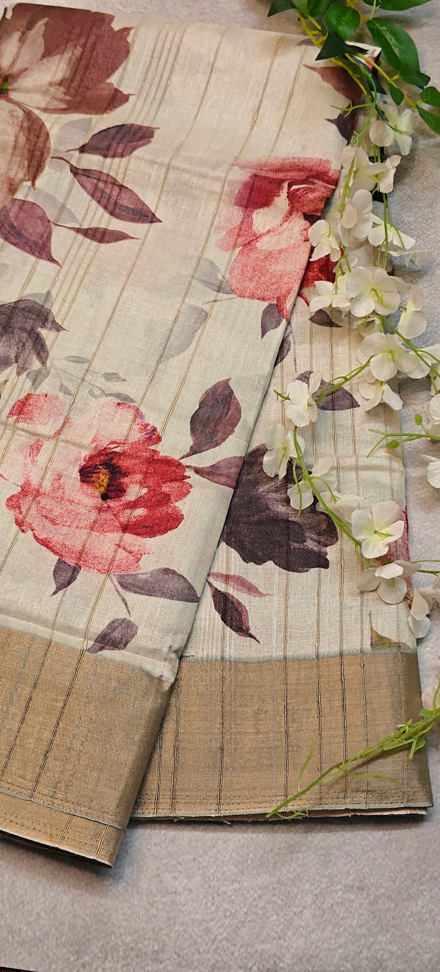 White blended silk saree with Kalamkari-inspired floral digital print in red, pink, and maroon tones with golden border lying flat and folded on a textured surface with green leaves and flowers.