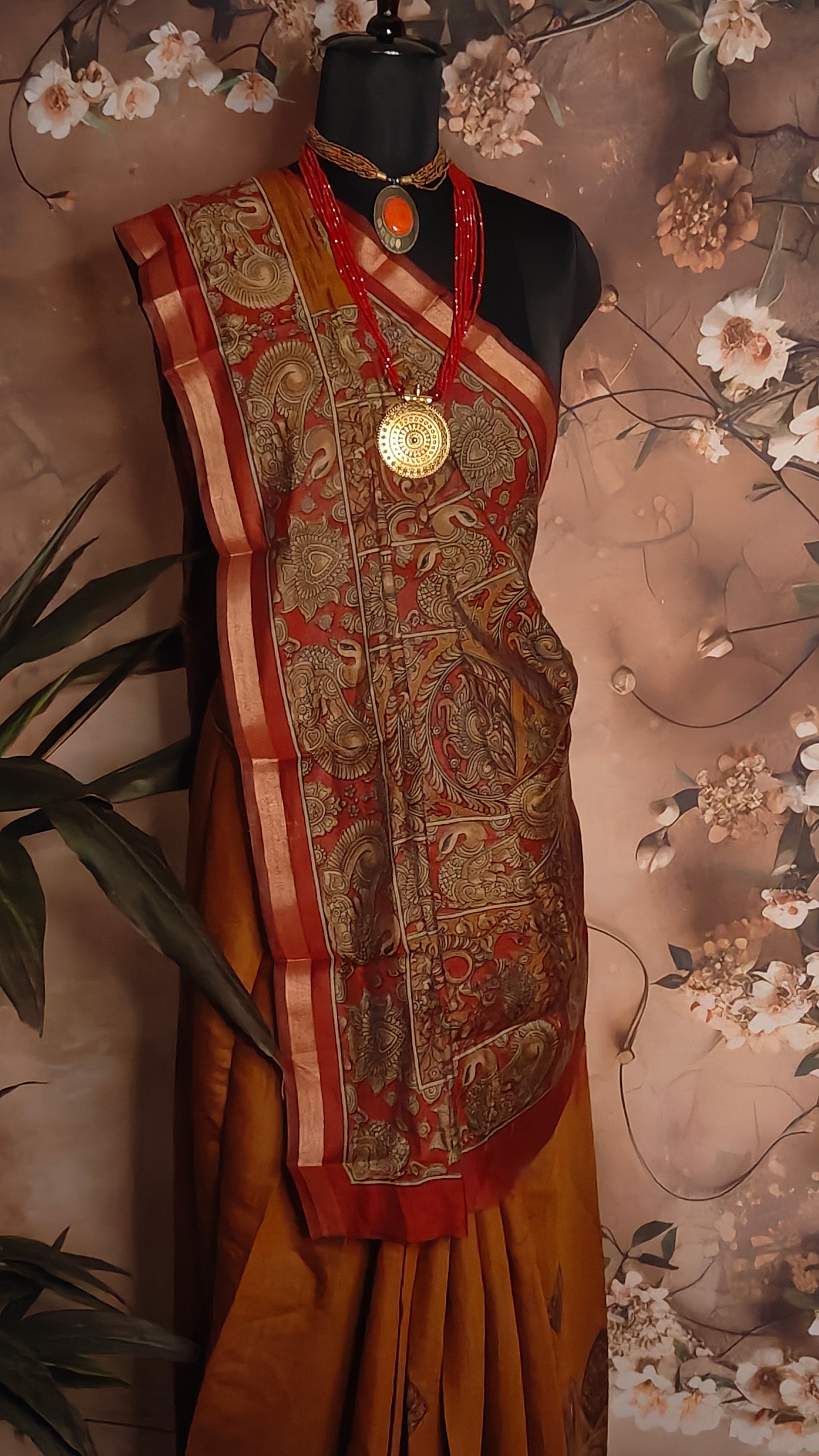 Maroon blended silk saree with Kalamkari-inspired floral and paisley digital print and broad golden border on a mannequin against a floral background.