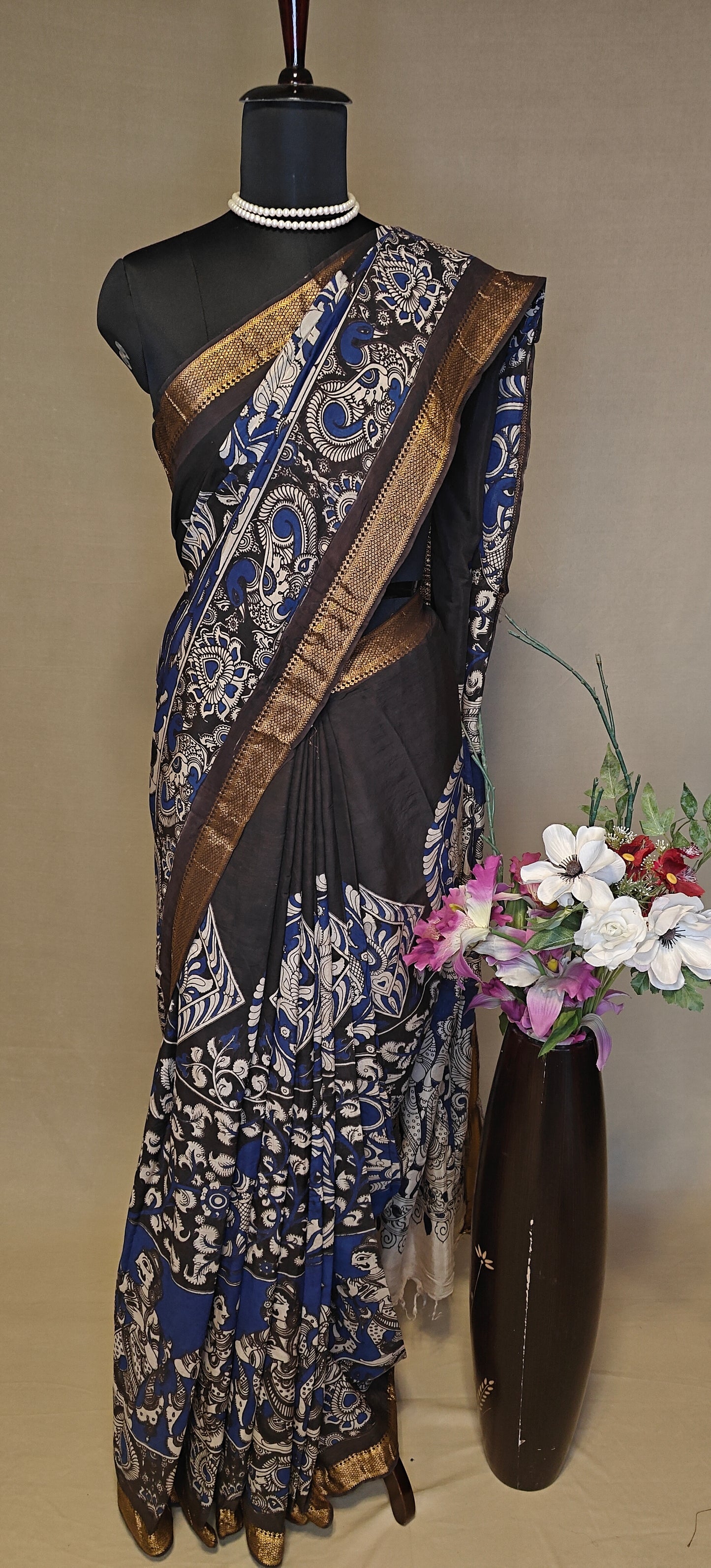 Black silk saree with Kalamkari-style floral and paisley print and golden border on a mannequin wearing a pearl necklace, against a plain background.