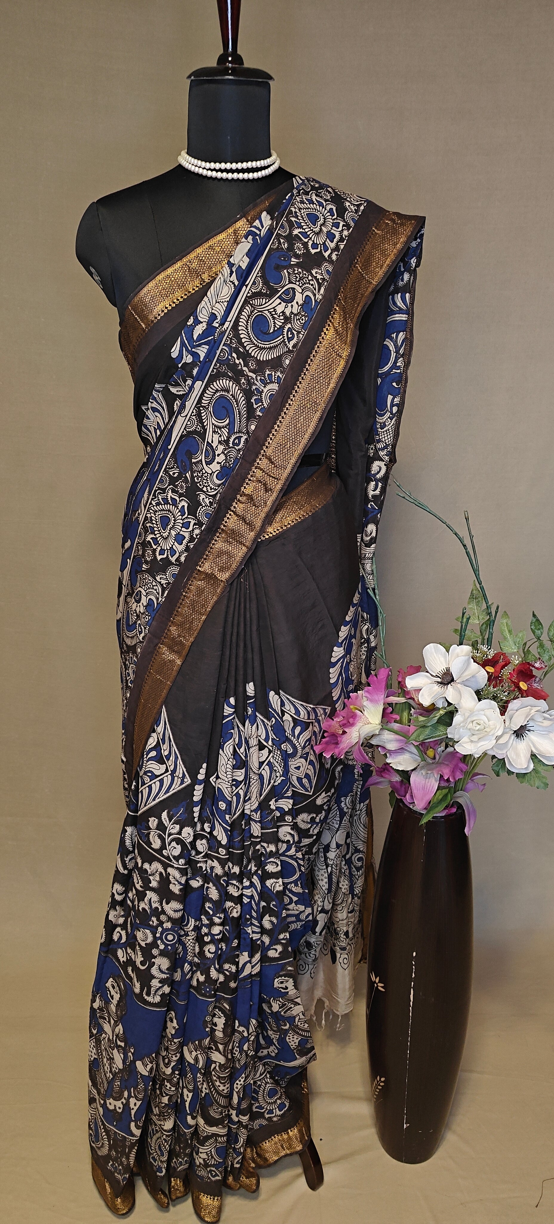 Black silk saree with Kalamkari-style floral and paisley print and golden border on a mannequin wearing a pearl necklace, against a plain background.