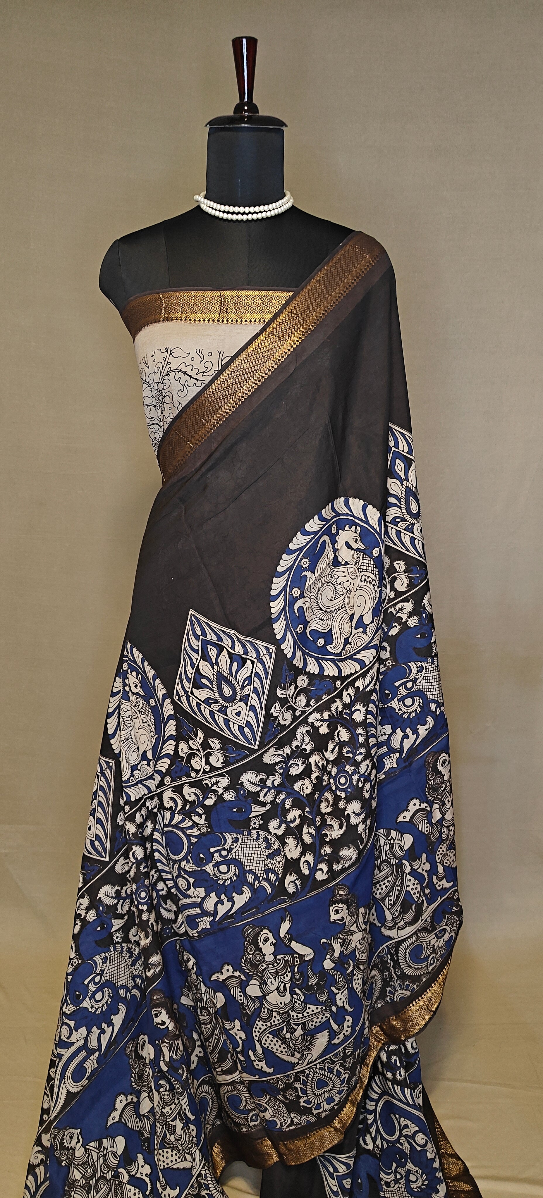 Black silk saree with Kalamkari-style floral and paisley print and golden border on a mannequin wearing a pearl necklace, against a plain background.