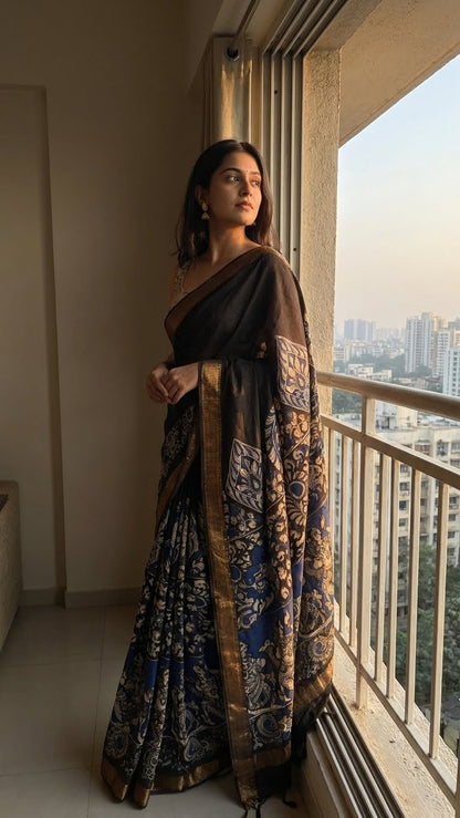 Woman in a black silk saree with kalamkari-style floral and paisley print and golden border standing by a window with a cityscape view.