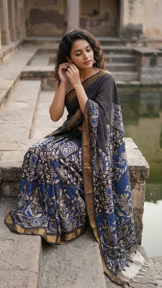 Woman in a black silk saree with kalamkari-style floral and paisley print and golden border sitting by a water body.
