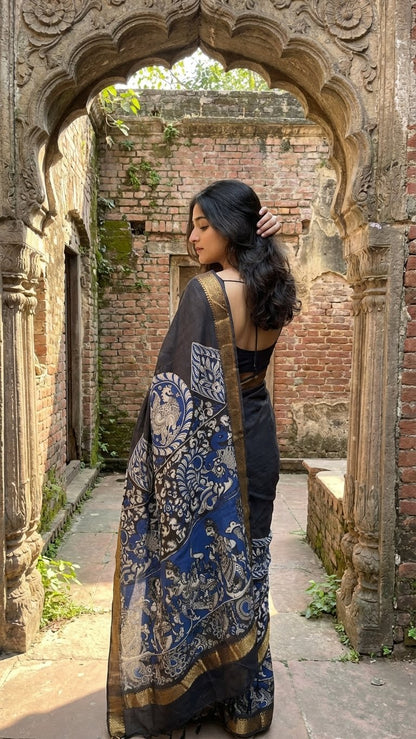 Woman in a black silk saree with kalamkari-style floral and paisley print and golden border standing in an archway of an old building.
