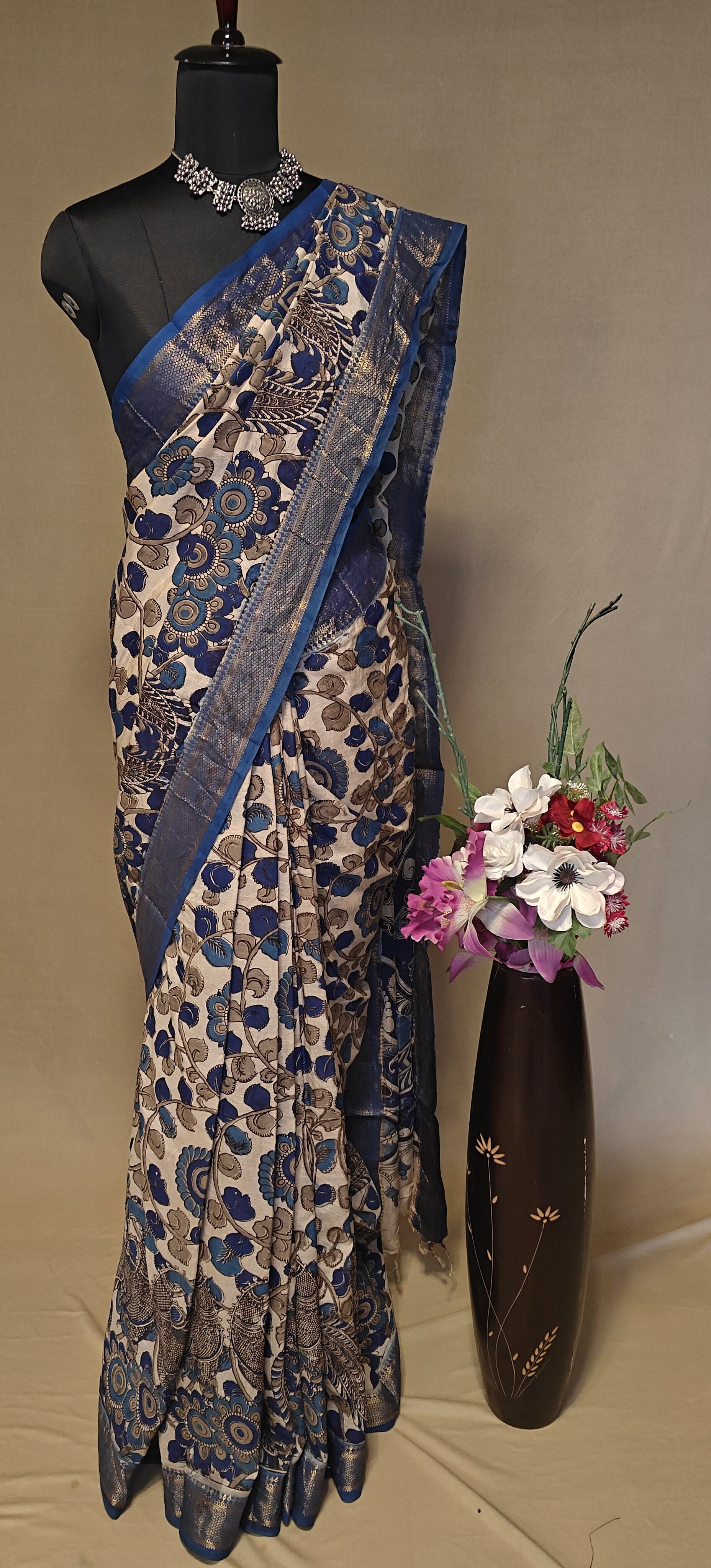 Indigo silk saree with Kalamkari-inspired digital print and golden border on a mannequin, next to a vase with flowers on a plain background.