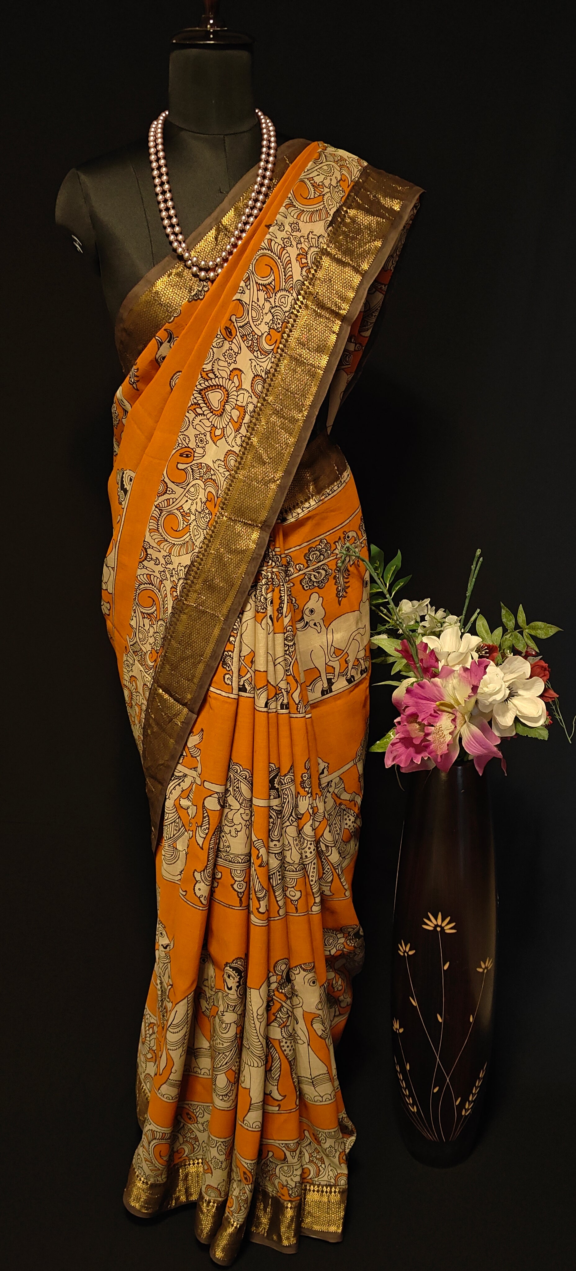 Orange silk saree with Kalamkari-style floral and paisley print and golden Mangalgiri border on a mannequin wearing a pearl necklace, against black background.