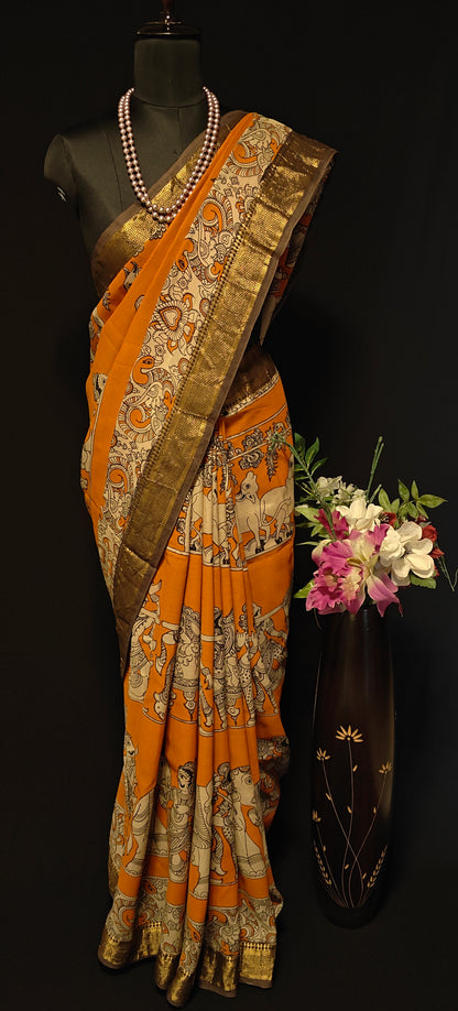 Orange silk saree with Kalamkari-style floral and paisley print and golden Mangalgiri border on a mannequin wearing a pearl necklace, against black background.