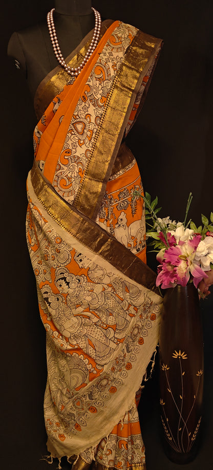 Orange silk saree with Kalamkari-style floral and paisley print and golden Mangalgiri border on a mannequin wearing a pearl necklace and showcasing the saree pallu with floral and paisley print, against black background.