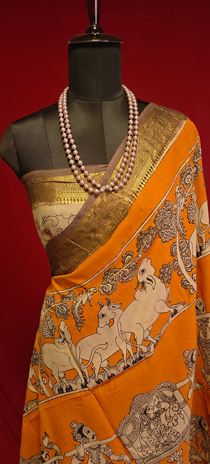 Orange silk saree with Kalamkari-style floral and paisley print and golden Mangalgiri border on a mannequin wearing a pearl necklace showcasing the blouse piece, against black background.