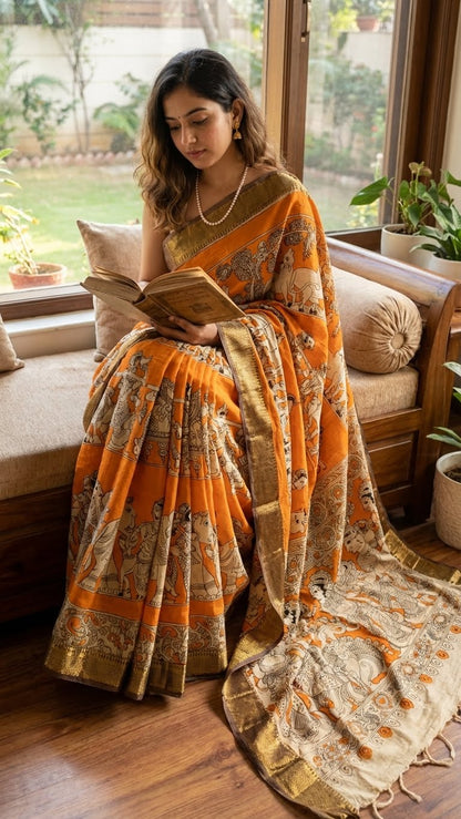 Woman in an orange silk saree with kalamkari-style floral and paisley print and golden mangalgiri border reading a book in a cozy room with large windows.