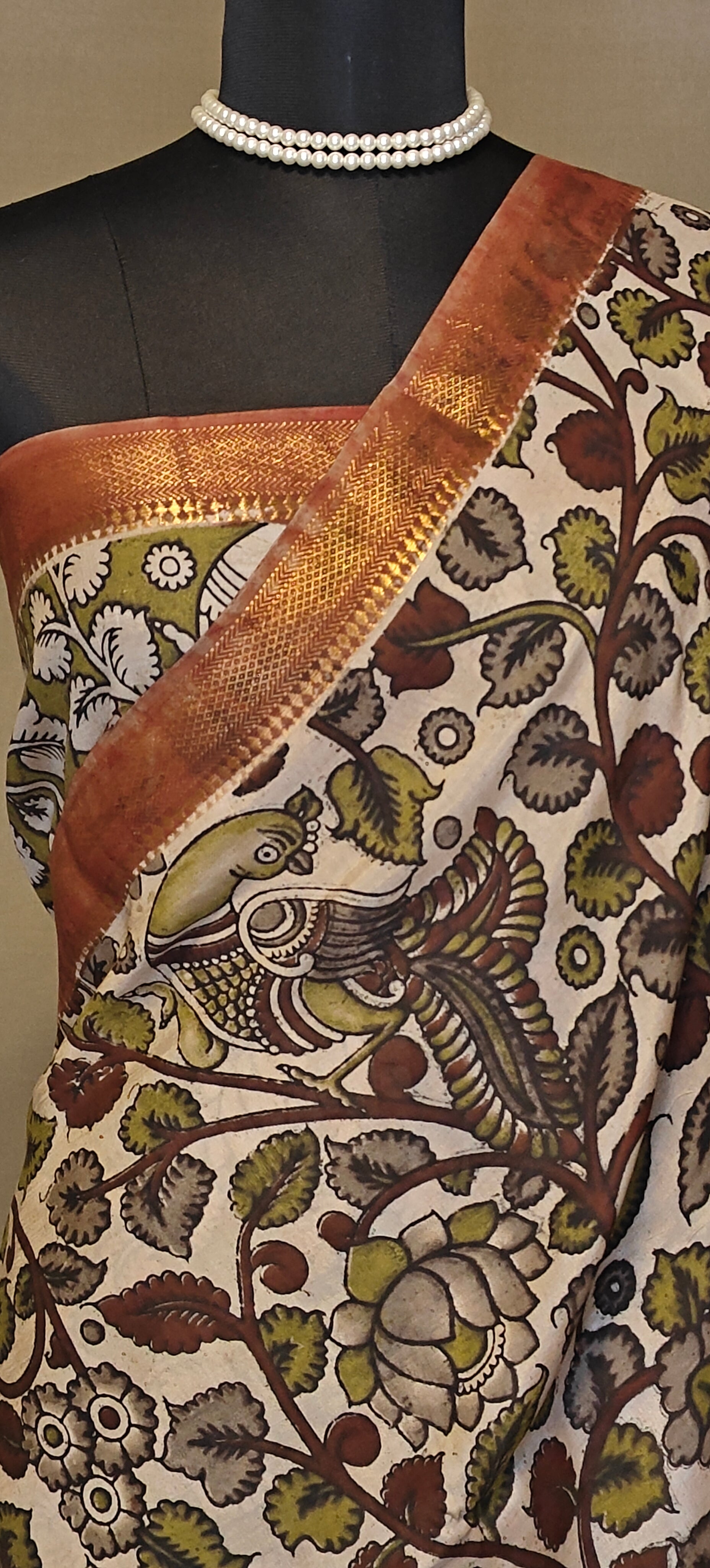 Brown silk saree with Kalamkari-inspired motifs and subtle golden border on a mannequin wearing a pearl necklace and blouse piece, against a neutral background.