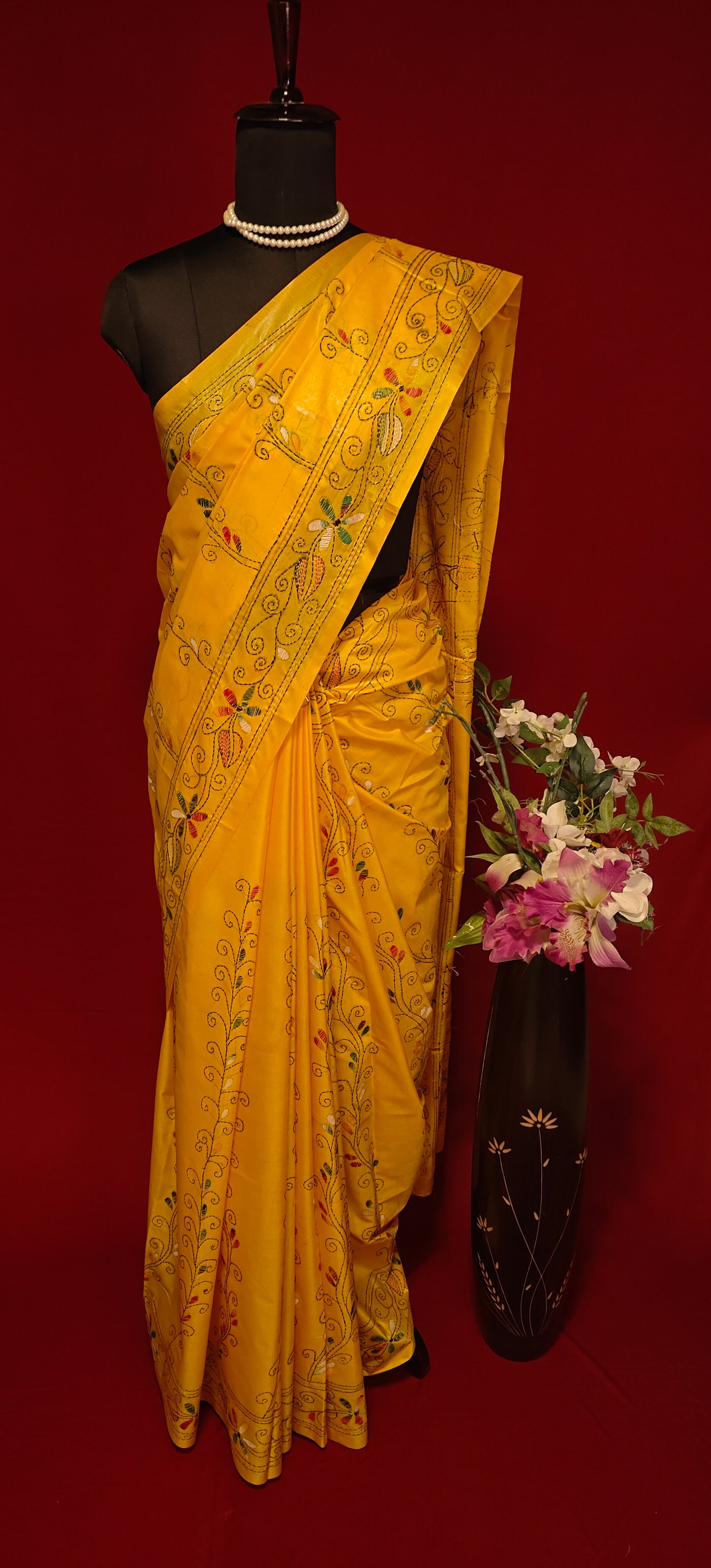 Handcrafted Kantha stitched cotton saree in yellow with multicolor floral and vine embroidery with flower vase on a mannequin against a red background.