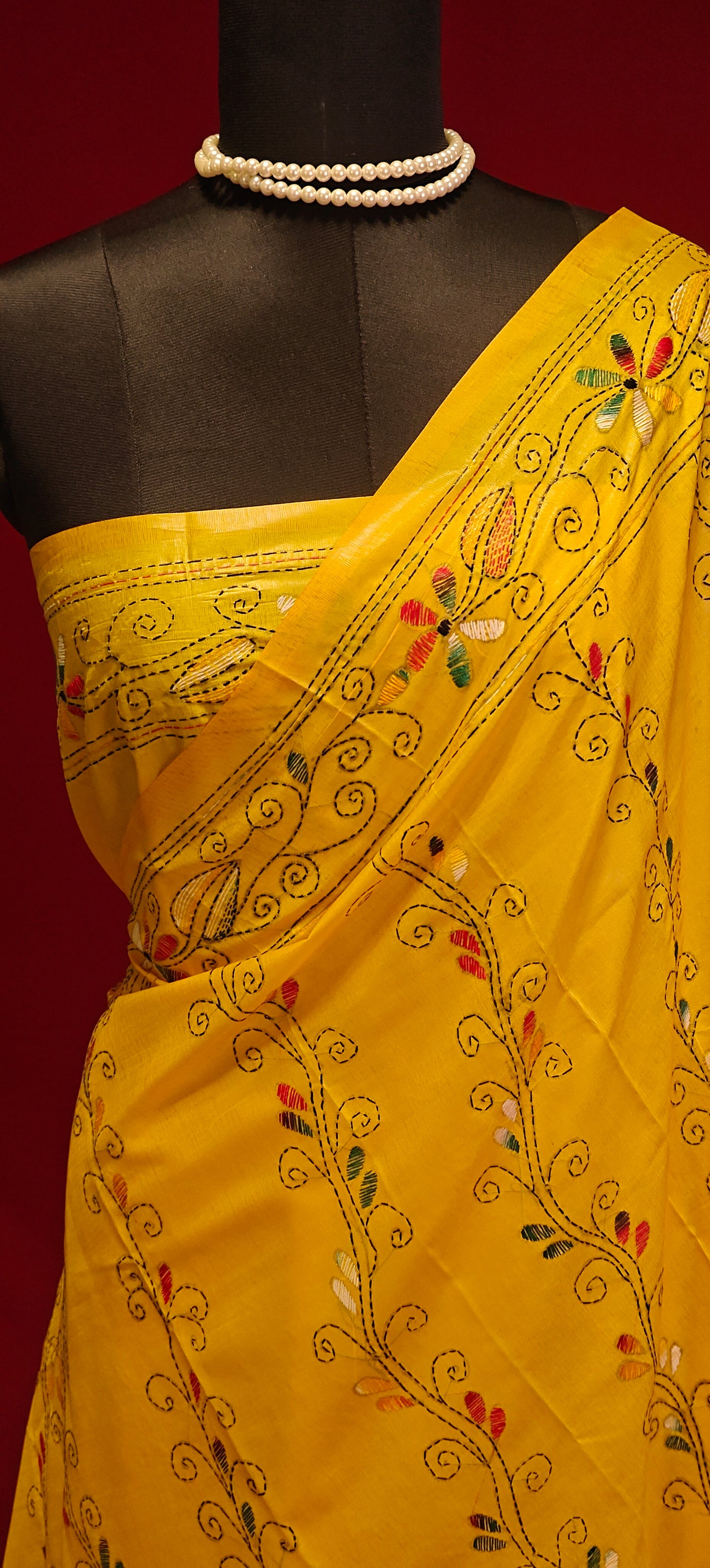 Handcrafted Kantha stitched cotton saree in yellow with multicolor floral and vine embroidery on a mannequin showcasing the blouse piece against a red background.