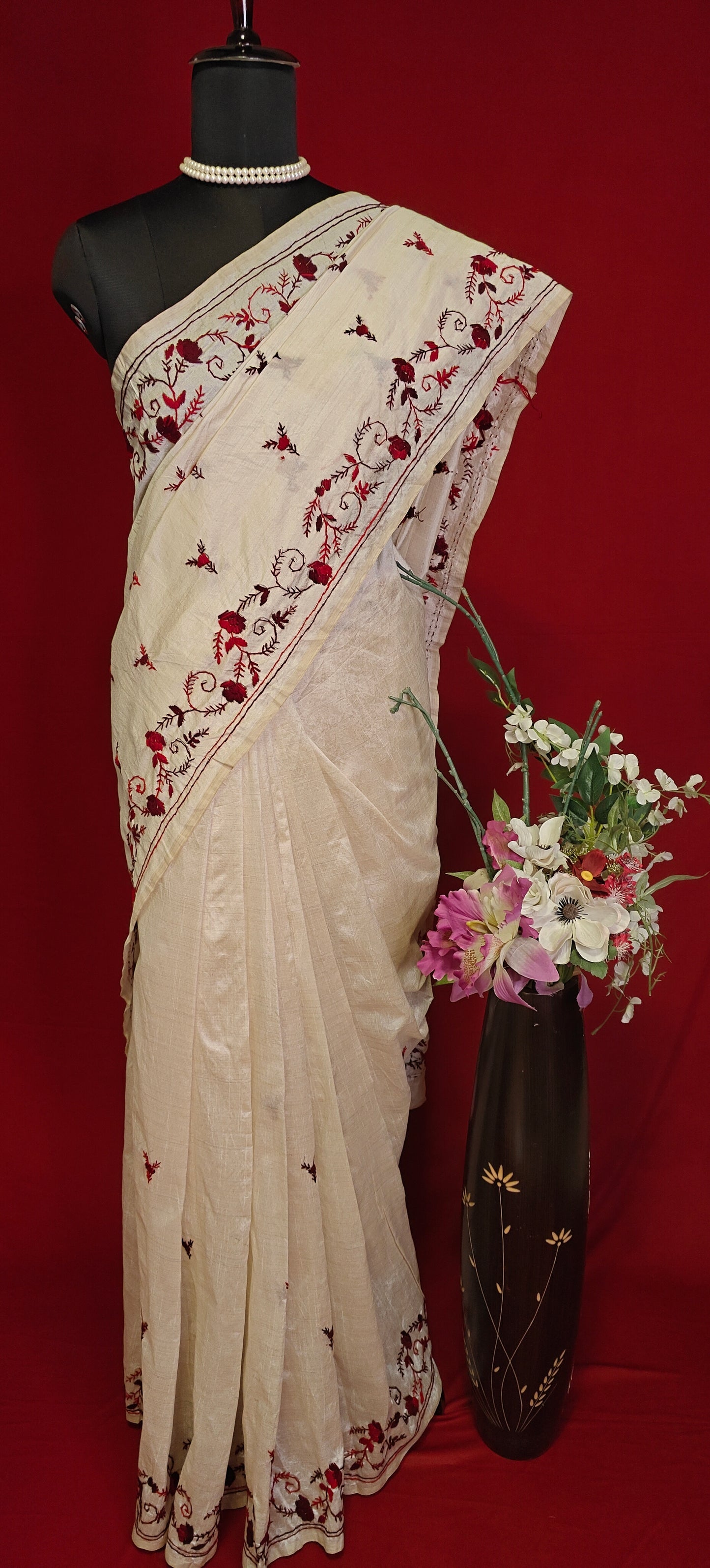 Handcrafted kantha stitched saree in cream with floral embroidery in maroon and red on a mannequin with a flower vase against a red background.