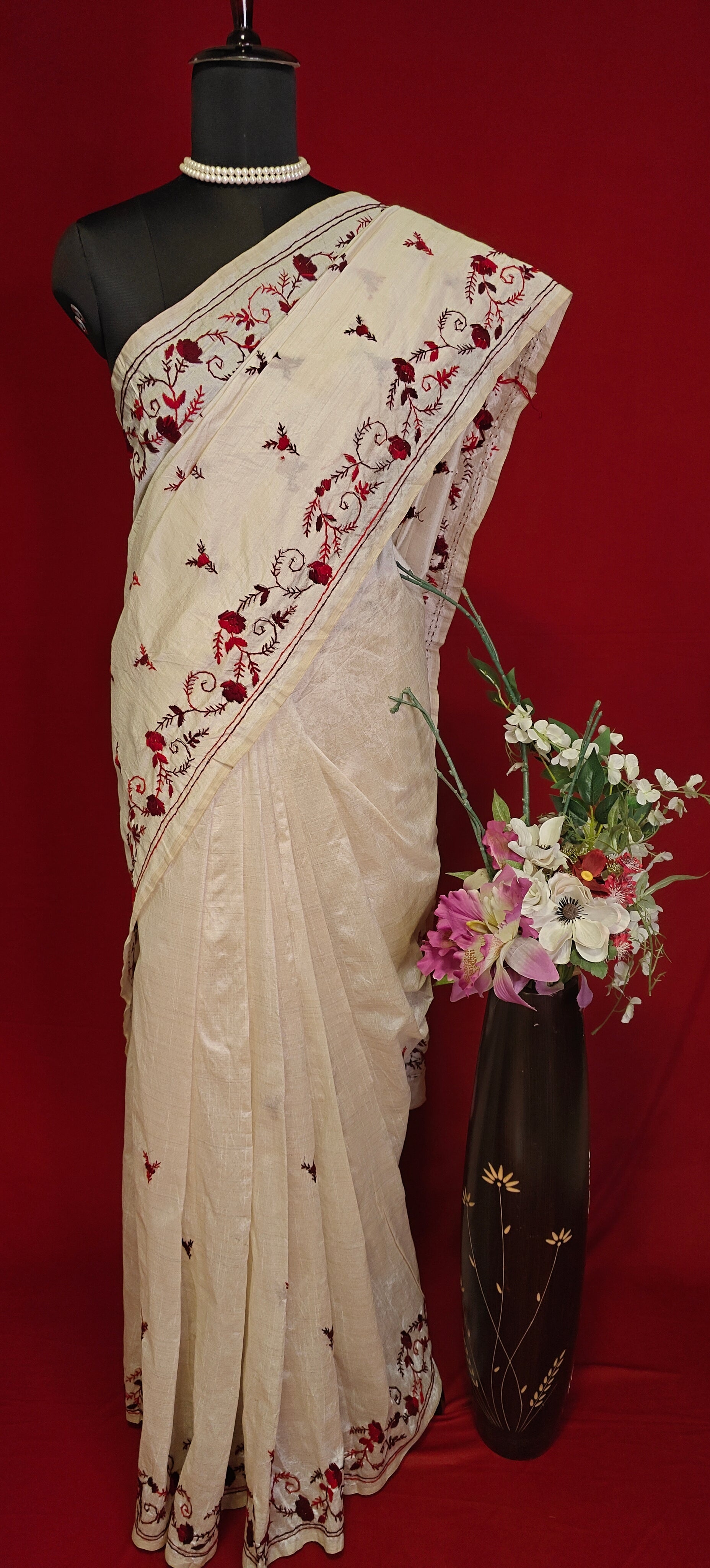 Handcrafted kantha stitched saree in cream with floral embroidery in maroon and red on a mannequin with a flower vase against a red background.